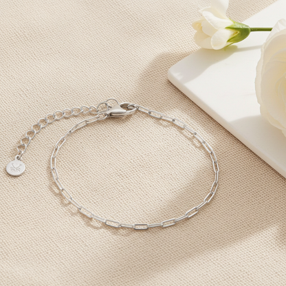 Silver paperclip bracelet on a beige surface with a white flower in the background